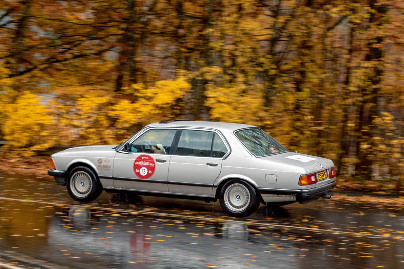 BMW E9 Coupé rijdt op een weg in de door een herfstig bos