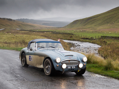 Austin Healey 3000 MK3 rijd op een weg in een drassig heuvellandschap